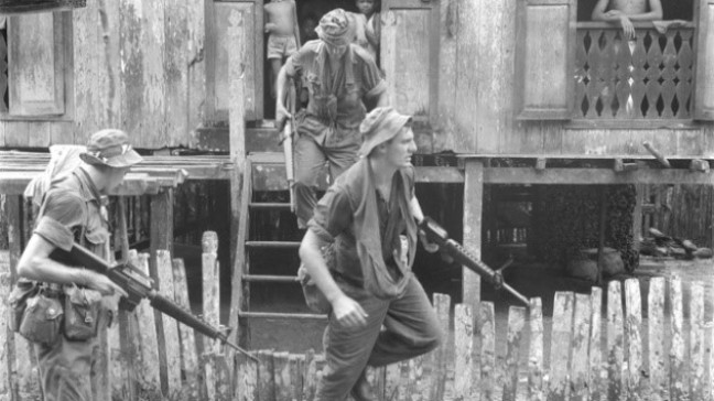 Soldiers from the 4th Battalion, The Royal Australian Regiment, moving from hut to hut in a village as they carry out a routine patrol along the Malaysian-Indonesian border. Left to right: Corporal John Maloney, from Coburg, Victoria; Private (Pte) Bill McBride, of Albany, WA, on the steps of the hut; and Pte Arthur Francis, of Cessnock, NSW. A number of children are watching them from inside the hut.