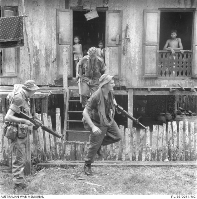 Soldiers from the 4th Battalion, The Royal Australian Regiment, moving from hut to hut in a village as they carry out a routine patrol along the Malaysian-Indonesian border. Left to right: Corporal John Maloney, from Coburg, Victoria; Private (Pte) Bill McBride, of Albany, WA, on the steps of the hut; and Pte Arthur Francis, of Cessnock, NSW. A number of children are watching them from inside the hut.